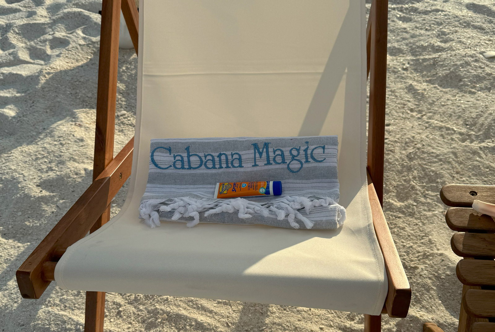 Home 15 Cabana Magic branded Turkish beach towel and sunscreen resting on a wood and canvas beach chair