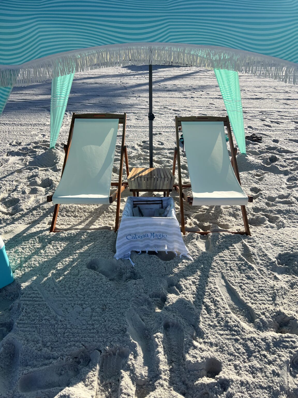 Home 3 Cabana Magic teal and white striped beach cabana with two canvas lounge chairs, a wooden beach table and a cooler draped with a branded Turkish towel on white sand